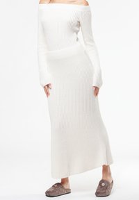 Robe en maille côtelée blanche, épaules dénudées, manches longues, coupe cintrée, avec des boutons décoratifs sur le devant ; associée à des chaussons marron.