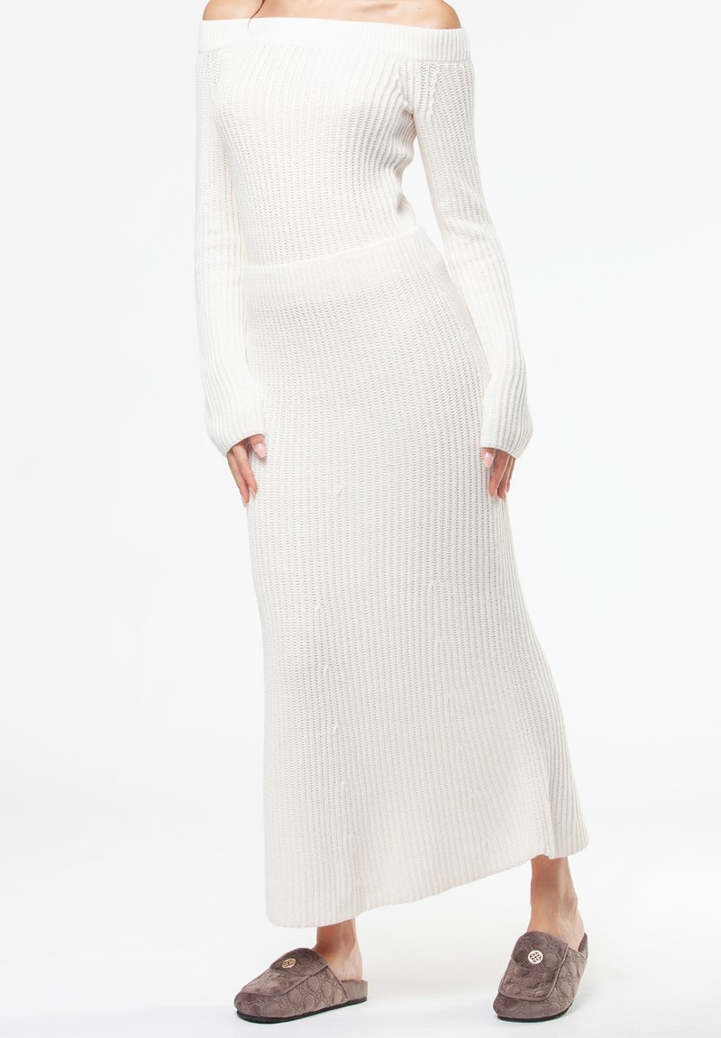 Robe en maille côtelée blanche, épaules dénudées, manches longues, coupe cintrée, avec des boutons décoratifs sur le devant ; associée à des chaussons marron.