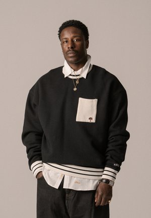 Svart hettegenser med hvite striper på ermene, med en kremfarget lomme og broderte trelogo. Laget over en hvit skjorte med krage.