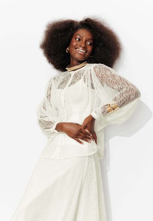 Femme souriante aux cheveux afro portant une robe en dentelle blanche et des bijoux en or, posant avec les mains sur les hanches devant un fond blanc.