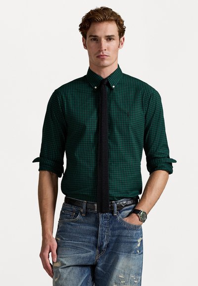 Chemise à carreaux verts avec col boutons, associée à une cravate noire et un jean en denim déchiré. Présente des manches retroussées et une montre au poignet.