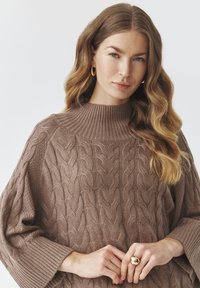 Brauner Kabelstrickpullover mit einem gerippten hohen Kragen und übergroßen Ärmeln, der ein strukturiertes Muster und ein weiches, gewebtes Material aufweist.