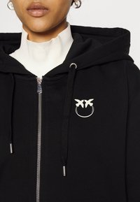 Femme portant un sweat à capuche noir zippé avec cordons et logo papillon blanc sur la poitrine, superposé à un haut blanc à col montant.