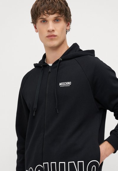 Fekete, cipzáras kapucnis pulóver, puha anyagból készült, elülső zsebekkel és fehér logóval a bal mellen és az alján.