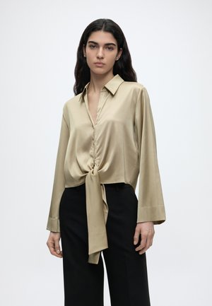 Frau mit langen dunklen Haaren trägt eine beige Satinkbluse, die an der Taille gebunden ist, und schwarze hoch taillierte Hosen vor einem schlichten weißen Hintergrund.