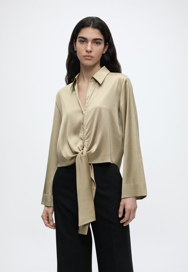 Γυναίκα με μακριά σκουρόχρωμα μαλλιά φοράει μπεζ σατέν μπλούζα δεμένη στη μέση και μαύρο παντελόνι με ψηλή μέση σε απλό λευκό φόντο.