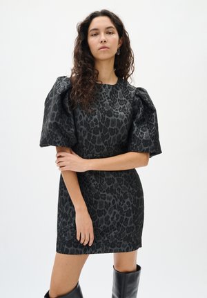 Frau mit langen lockigen Haaren, die ein dunkelgraues Leopardenmuster-Kleid mit Puffärmeln und schwarzen kniehohen Stiefeln trägt, posiert vor einem weißen Hintergrund.