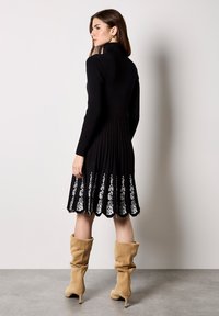 Zwarte geribbelde jurk met lange mouwen en een hoge kraag, voorzien van witte bloemborduursels aan de zoom en een uitlopende rokontwerp.