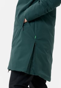 Lange, donkergroene waterdichte jas met een ritsventilatie aan de zijkant en een handzak. Lichtjes gestructureerde stof met een strakke, minimalistische uitstraling.