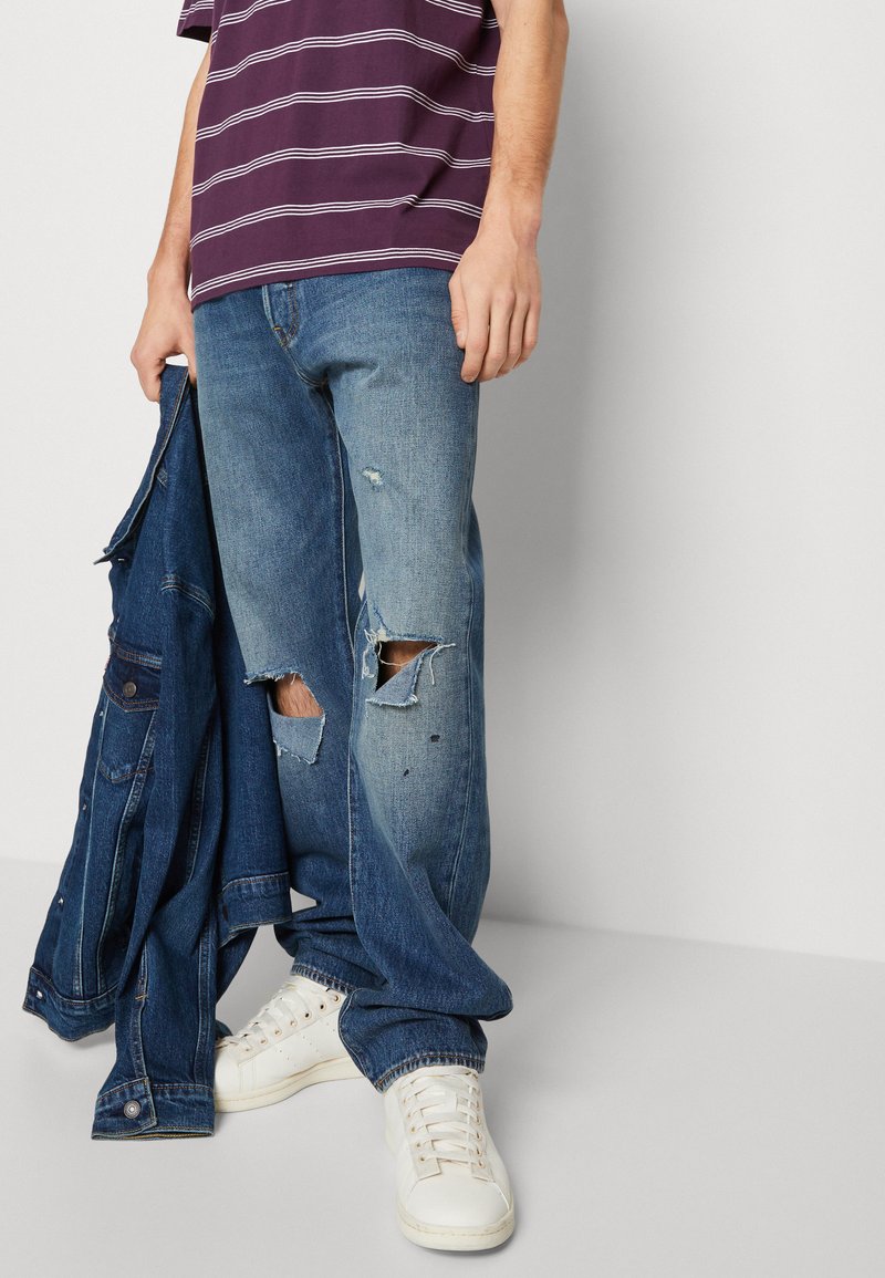 Homme portant un jean bleu déchiré, des baskets blanches et une chemise rayée violette, tenant une veste en denim bleu foncé à ses côtés.