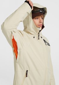 Beige waterdichte jas met capuchon, voorzien van oranje mesh accenten, ritszakken en een logo op de borst. Soepele textuur.
