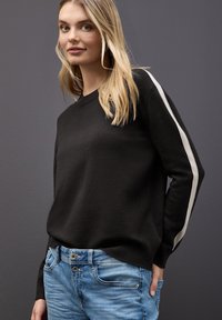 Czarny sweter z okrągłym dekoltem, z białymi paskami na rękawach, w połączeniu z jasnoniebieskimi dżinsami. Miękka tekstura, luźny krój.