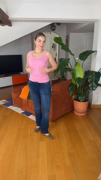 Νεαρή γυναίκα με ροζ αμάνικο μπλουζάκι και τζιν, στέκεται στο σαλόνι κοντά σε μεγάλο φυτευμένο φυτό και καφέ καναπέ με πορτοκαλί κουβέρτα.