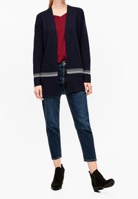 Marinblå cardigan med öppen framsida och randiga detaljer vid ärmsluten, ihopparad med en röd topp och mörkblå jeans. Texturen ser mjuk ut.