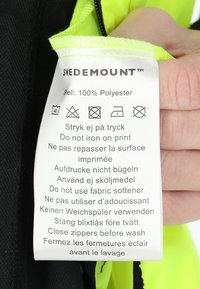Etykieta na kurtkę wykonaną w 100% z poliestru, zawierająca instrukcje dotyczące pielęgnacji w kilku językach. Kontrast kolorystyczny między metką a jaskrawożółtym materiałem.