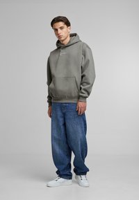 Felpa grigia con cappuccio e tasca frontale, logo bianco, abbinata a jeans blu larghi e sneakers bianche, su uno sfondo neutro.