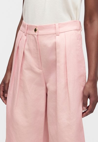 Gros plan d'une personne portant un pantalon plissé rose clair taille haute avec un haut blanc, montrant le bouton devant et les passants de ceinture.