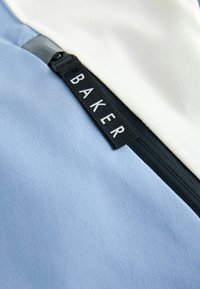 Närbild av en svart dragkedja med texten "BAKER" på ljusblå och vit texturerad tyg.