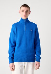 Blauer Strickpullover mit hohem Reißverschlusskragen, gerippten Bündchen und Saum, versehen mit einem kleinen Logodetail auf der Brust.