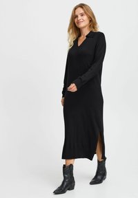 Vestido largo de punto negro con escote en V y aberturas laterales, combinado con botas de vaquero negras que presentan un detallado intrincado.