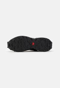 Black rubber sole with aggressive triangular lugs, a small red triangular detail near the center, designed for traction on rugged terrain.