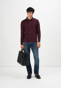 Muž v burgundském polo s dlouhým rukávem, modrých džínách a černých střevících s třásněmi, drží černý kožený aktovku před bílým pozadím.