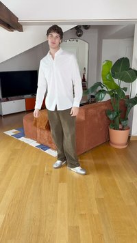 Chemise blanche à boutons, pantalon en suede vert olive et chaussures argentées. Canapé en cuir texturé marron dans une pièce avec un sol en bois et une plante en pot.