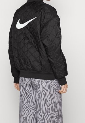 Personne portant une veste matelassée noire Nike avec un grand swoosh blanc dans le dos et un pantalon gris à rayures zébrées, vue de dos.