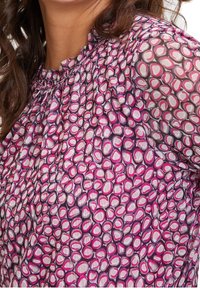 Vloeiende blouse van doorschijnende stof met een patroon van roze en witte afgeronde vormen op een paarse achtergrond; geplooide halslijn en mouwen.