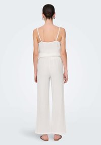 White sleeveless jumpsuit with a relaxed fit, wide legs, cinched waist, and thin adjustable straps. Texture appears soft and lightweight.
