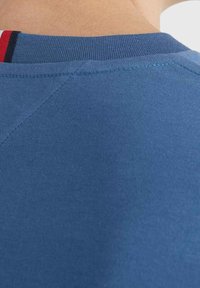 Camiseta de algodón azul con un cuello acanalado y una textura sutil. Se pueden ver detalles de bloques de color en rojo y negro en el borde del cuello.