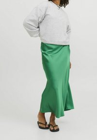 Felpa grigia, gonna midi in satin verde, texture liscia, design fluttuante e ciabatte nere. Collana a forma di cuore ben visibile.