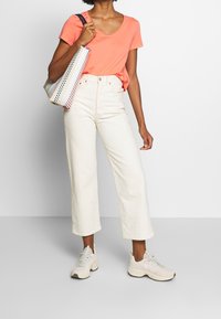 Korálové tričko s krátkým rukávem, bílé kalhoty s vysokým pasem a širokým střihem, světlé tenisky a taška s vícebarevným vzorem.