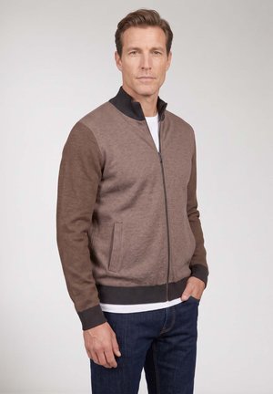 Mann trägt einen braunen Pullover mit Reißverschluss, dunklem Kragen und Bündchen, darunter ein weißes Hemd und blaue Jeans, steht vor neutralem Hintergrund.