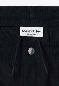 Pantaloni pantaloncini neri dal taglio rilassato con vita elasticizzata, cordoncino e accessori in metallo argentato. L'etichetta presenta il marchio "LACOSTE" e il logo del coccodrillo.