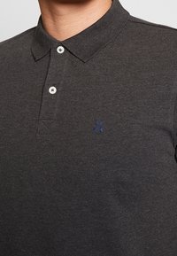 Polo gris foncé en tissu texturé avec un col, comportant deux boutons blancs et un petit logo bleu sur le côté gauche.