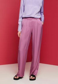 Růžové saténové kalhoty s volným střihem a záhyby, kombinované se světlým fialovým topem, na červeném pozadí. Na nohou černé sandály.