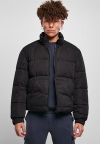 Chaqueta de plumas negra, diseño corto, con cuello alzado, cierre de cremallera en la parte delantera y puños elásticos. Textura suave y acolchada con bolsillos laterales.