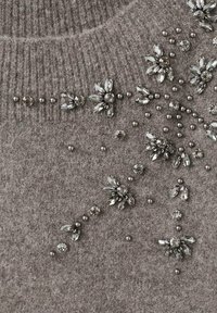 Suéter gris de punto con diseño floral adornado. Presenta cuentas metálicas y acentos de cristal en varias formas y tamaños.