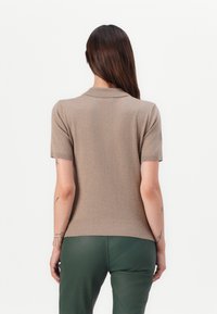 Kortermet, lys brun strikket topp med ribbestrikket krage og nederkant. Har en jevn tekstur og tettsittende design, kombinert med grønne bukser.
