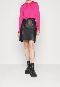 Fuchsia Bluse mit langen Ärmeln und Falten, kombiniert mit einem schwarzen Lederrock mit weißen Nähten und Knopfakzenten. Schwarze Plateauboots bis zu den Knöcheln.