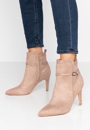 Bottines en suede beige clair avec un bout pointu, fermeture éclair et accents décoratifs perforés. Présente un talon haut et fin.