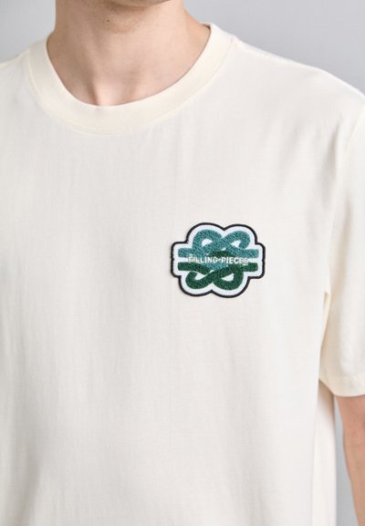Kreminės spalvos medvilninis marškinėliai su žaliai ir juodai siuvinėta logotipo etikete, ant kurios užrašyta "Filling Pieces", krūtinėje.