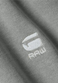 Šedá tkanina s texturovanou vazbou, na které je vyšitý bílý logo s nápisem "RAW" v moderním písmu.