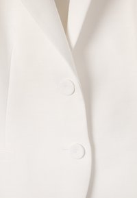 Gros plan de l’avant d’un blazer blanc montrant deux boutons, un revers et une partie de poche sur un tissu lisse.