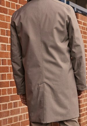 Abrigo marrón con cuadros, de corte relajado, mangas largas y detalle de abertura en la parte trasera, hecho de tela lisa sobre un fondo de ladrillos.