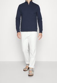 Felpa con zip color blu navy, pantaloni bianchi e sandali grigi. La felpa presenta un tessuto testurizzato e polsini a costine; i pantaloni hanno un taglio dritto.