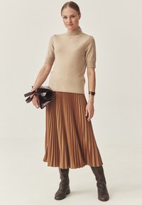 Maglione beige a maglia con maniche corte, abbinato a una gonna pieghettata marrone. Modella che regge una borsa a tracolla marrone scuro, indossando stivali marroni fino al ginocchio.