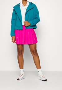 Teal huva jacka, vit polo, ljusrosa plisserad kjol med färgglada mönster och grå sneakers med röda accenter och vita strumpor.