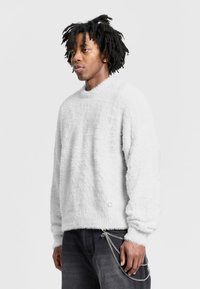 Fuzzy white sweater with a round neck and long sleeves, featuring a textured finish. Paired with dark jeans and a chain accessory.
